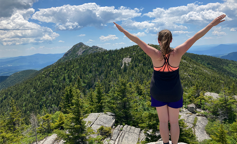 Woman stands on top of a mountain in the White Mountain National Forest. Trees are seen below.